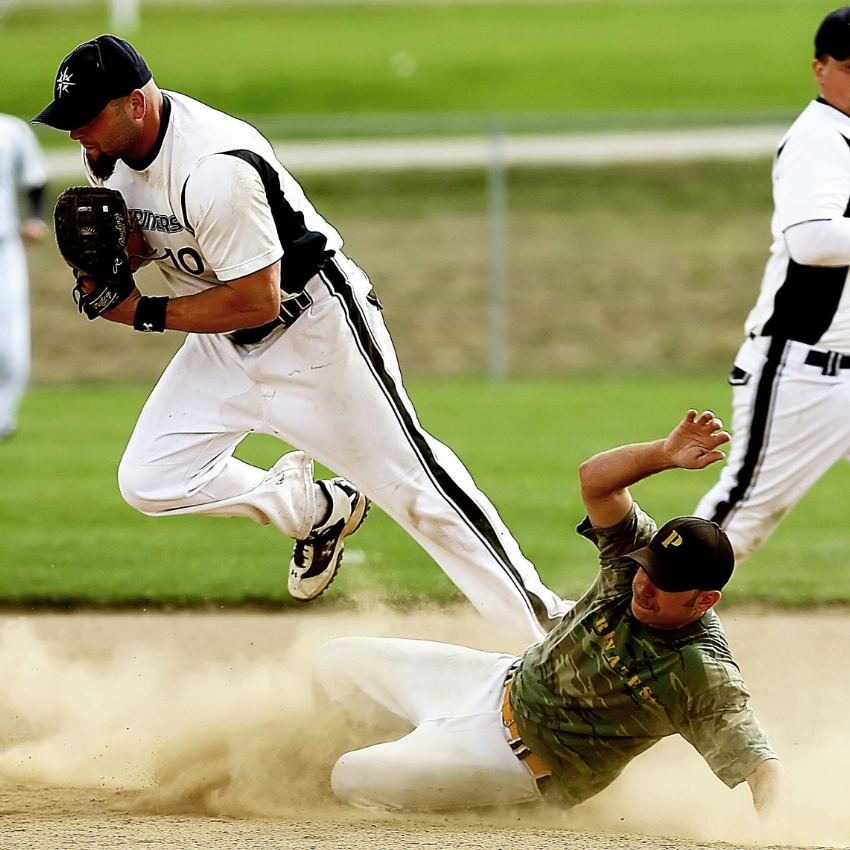 Baseball player in white uniform jumps over sliding opponent in camouflage uniform.