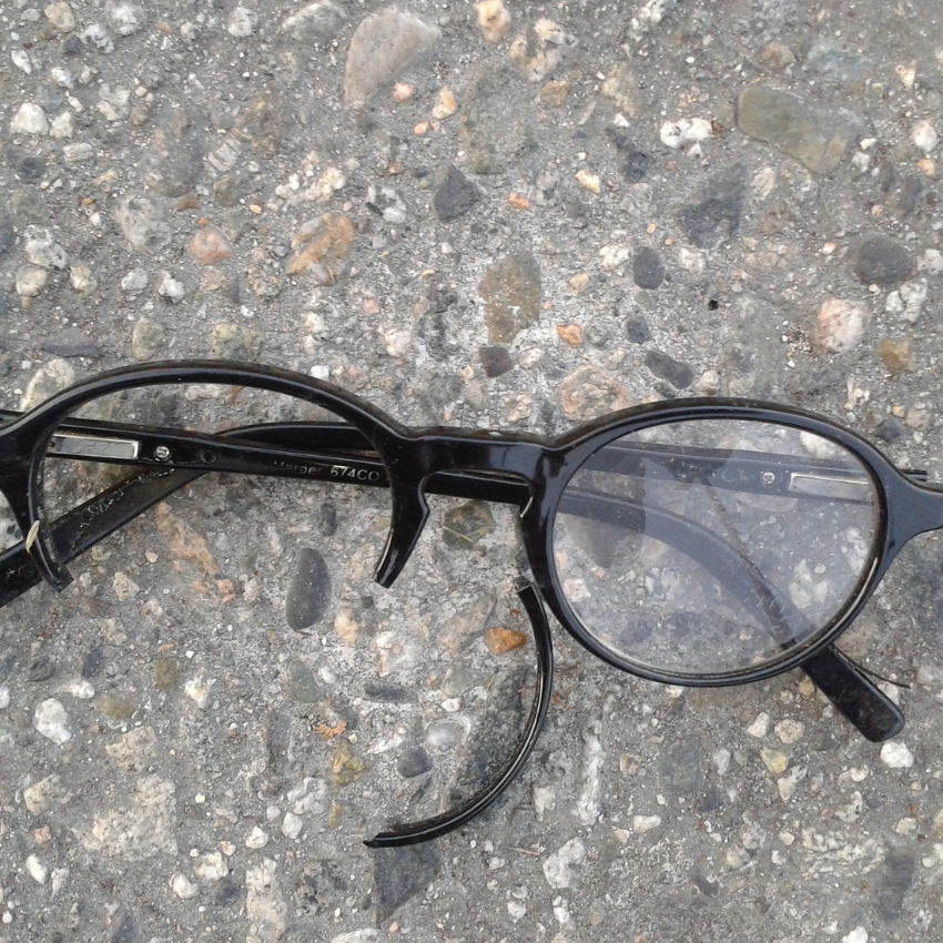 Broken black-rimmed eyeglasses on a rough, pebbled surface. Text on frame: "Harper 574CO."