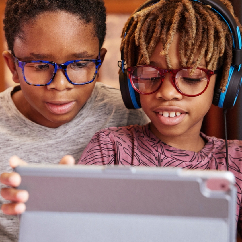 Two children wearing glasses, one with blue frames and one with red, using a tablet.