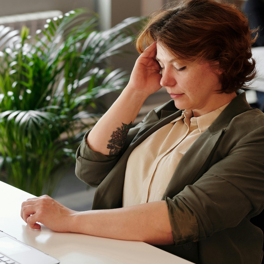 Woman in a green blazer sitting at a desk, holding her head, appearing stressed or tired.