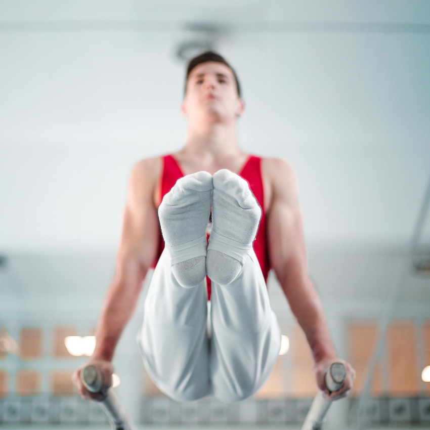 Gymnast in red tank top and white pants performing on parallel bars, focus on feet and hands.