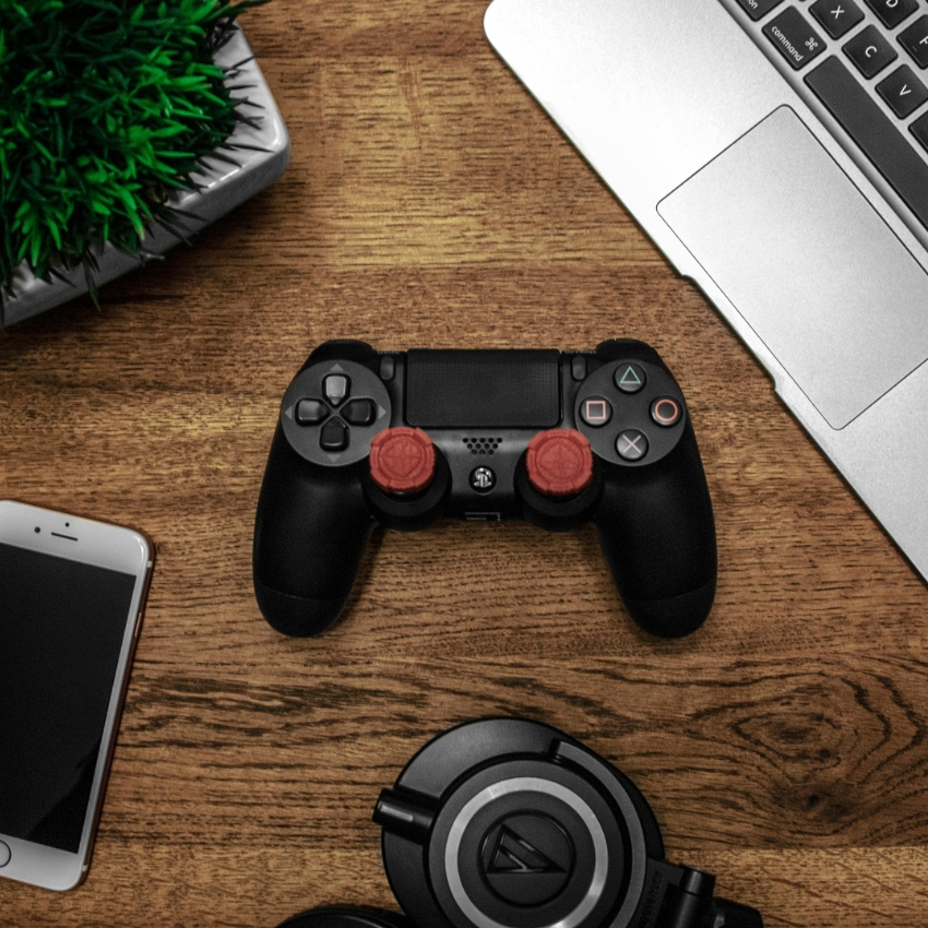 Black gaming controller with two red thumb grips, displayed on a wooden surface near a laptop, smartphone, and headphones.