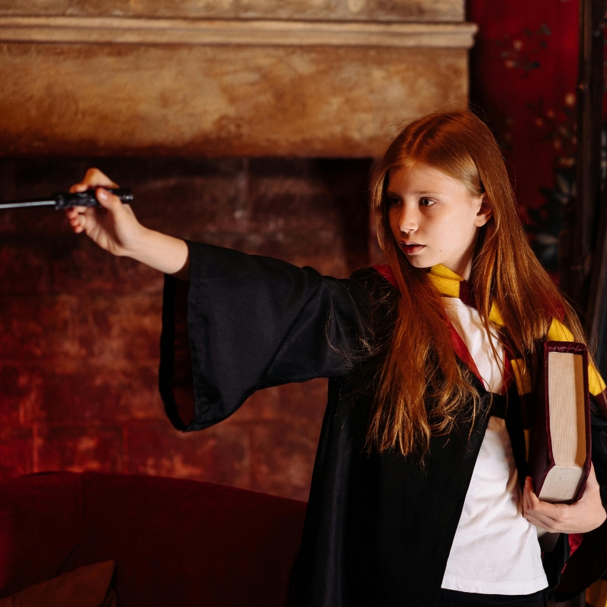 Girl in a black robe holding a wand and book, wearing a red and yellow striped scarf.