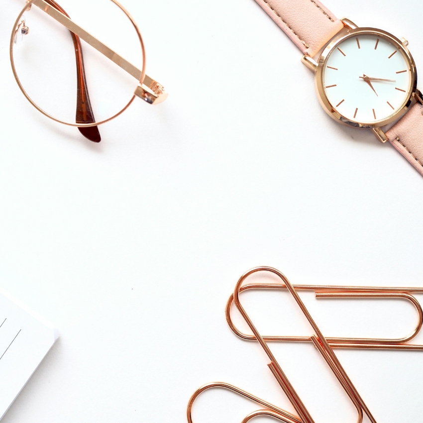 Gold-rimmed eyeglasses, a rose gold watch with a pink strap, and large gold paper clips on a white surface.