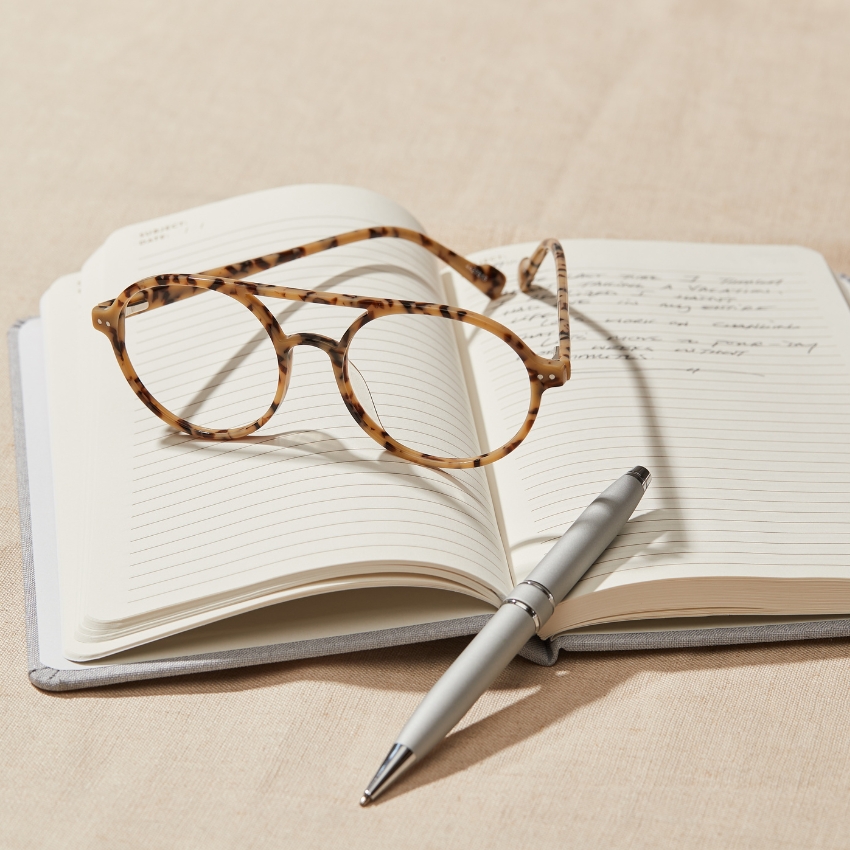 Tortoiseshell glasses resting on an open notebook with a silver pen. Text in the notebook is cursive handwriting.