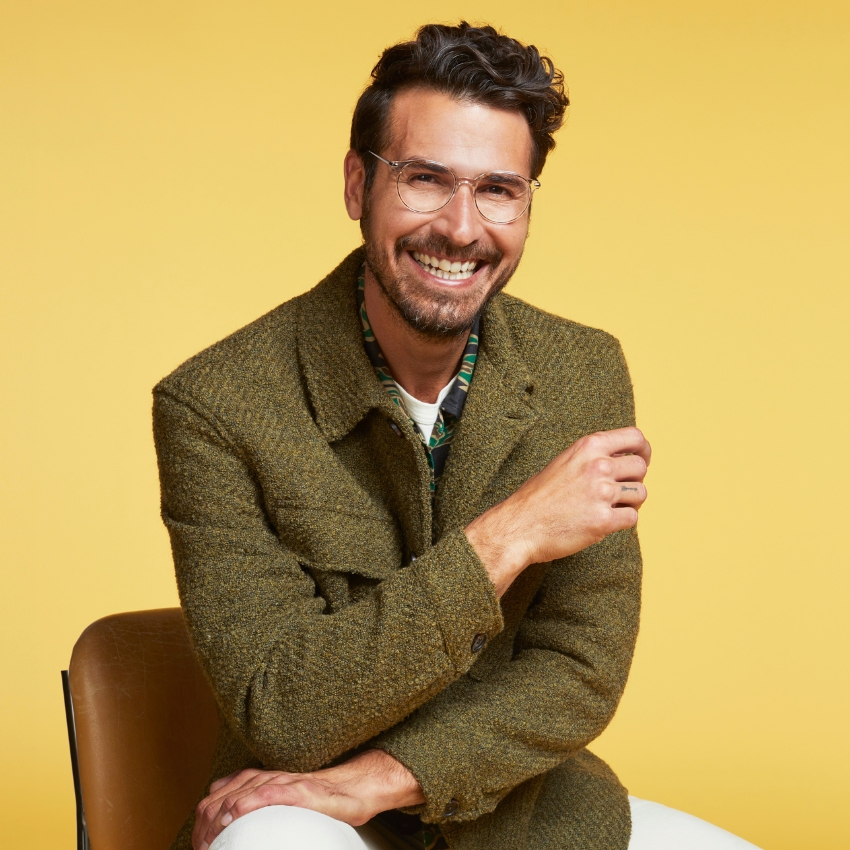 Man in a green wool coat and clear glasses sitting on a chair with a yellow background.