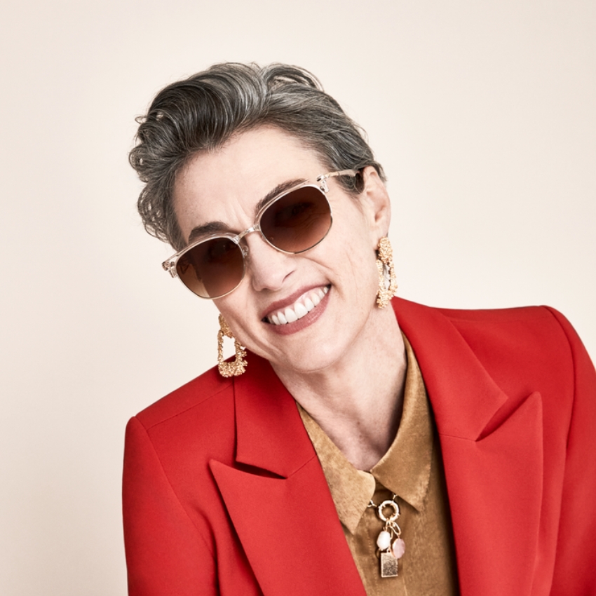 Woman wearing sunglasses, gold earrings, red blazer, and tan shirt with a unique pendant necklace.