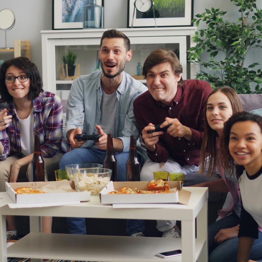 Group enjoying video games with pizza and snacks on the table, including beer bottles and popcorn bowls.