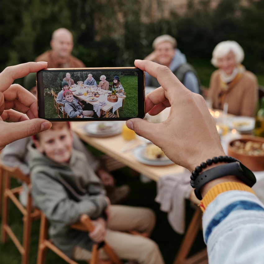 Hands holding a smartphone taking a photo of a family gathered around a table outdoors.