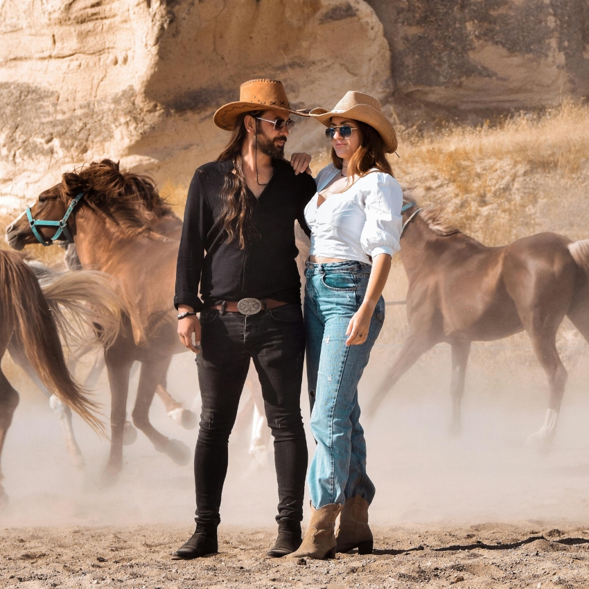 Two people wearing cowboy hats and sunglasses stand in front of galloping horses.