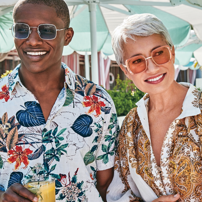 Two people wearing colorful, patterned button-up shirts and sunglasses smiling.