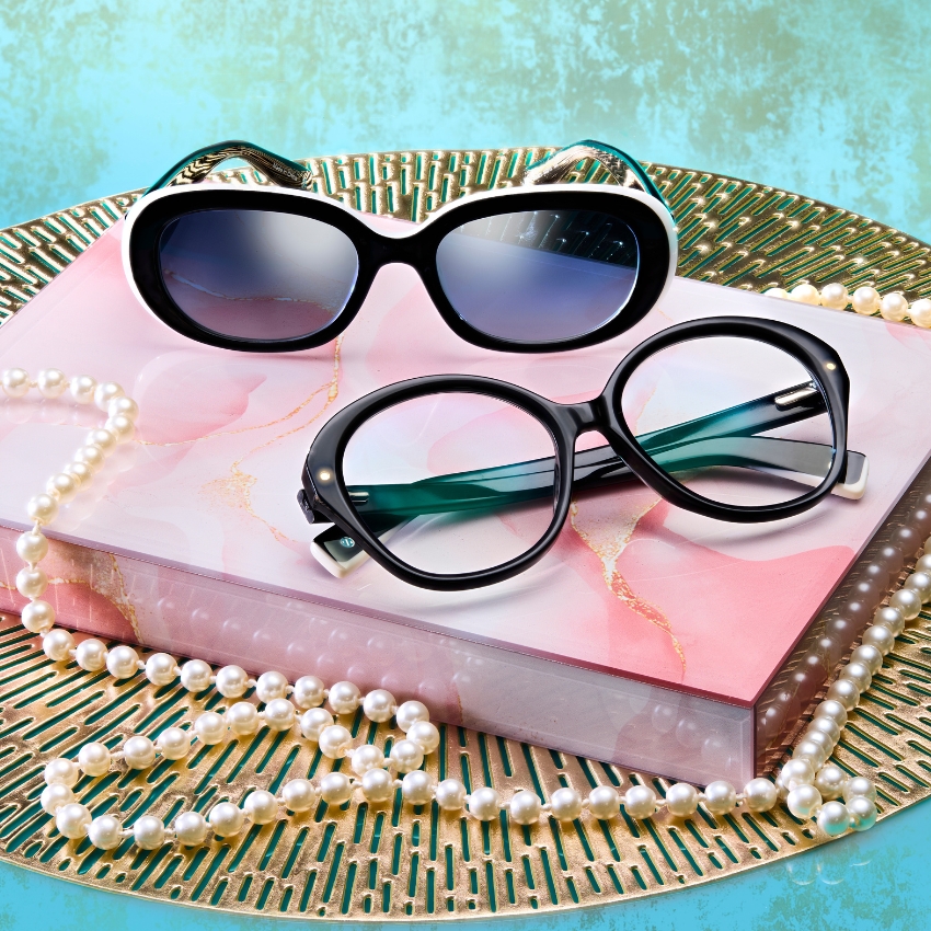 Two pairs of black-framed glasses sit on a pink book surrounded by pearl necklaces.