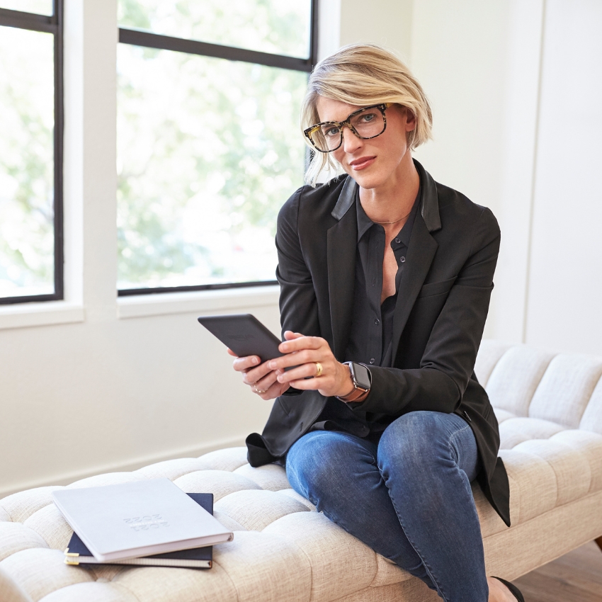 Woman in black blazer and blue jeans holding a tablet, with notebooks beside her.