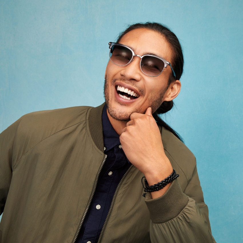 Man wearing aviator-style sunglasses, green bomber jacket, and black braided bracelet, smiling broadly.