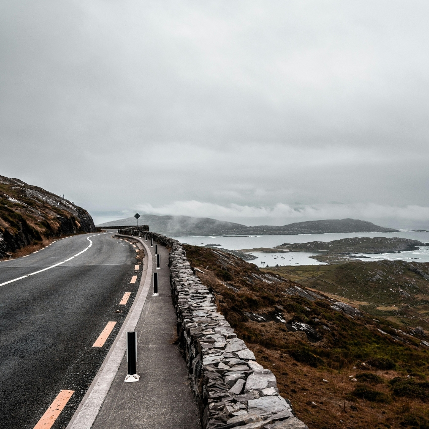 Winding road along a stone wall overlooking a misty, coastal landscape with hills and water in the distance.