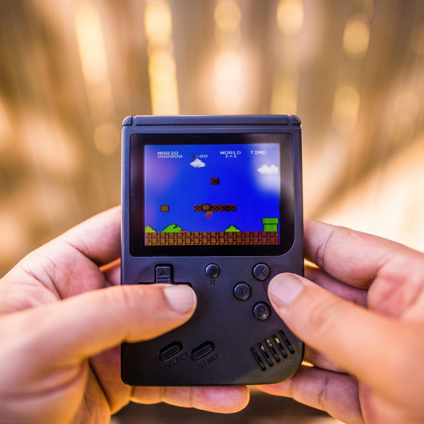 Hands holding a black handheld gaming console with the game Super Mario displayed on the screen. Text on screen: "MARIO 000000 WORLD TIME"