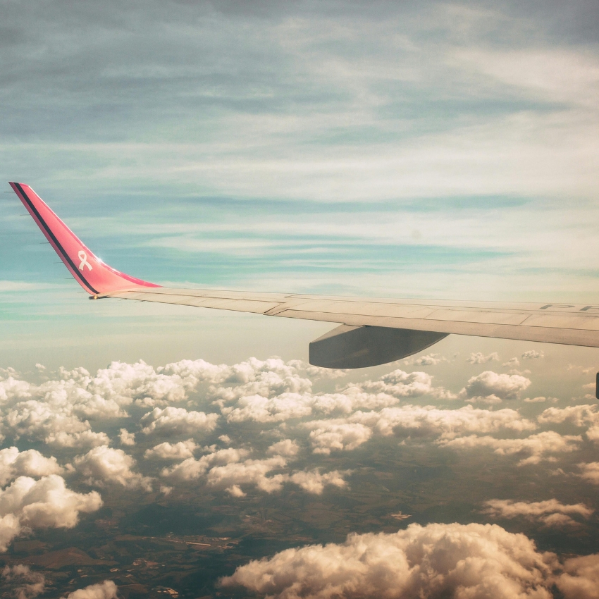 Airplane wing with a pink ribbon on the wingtip, flying above clouds under a partly cloudy sky.