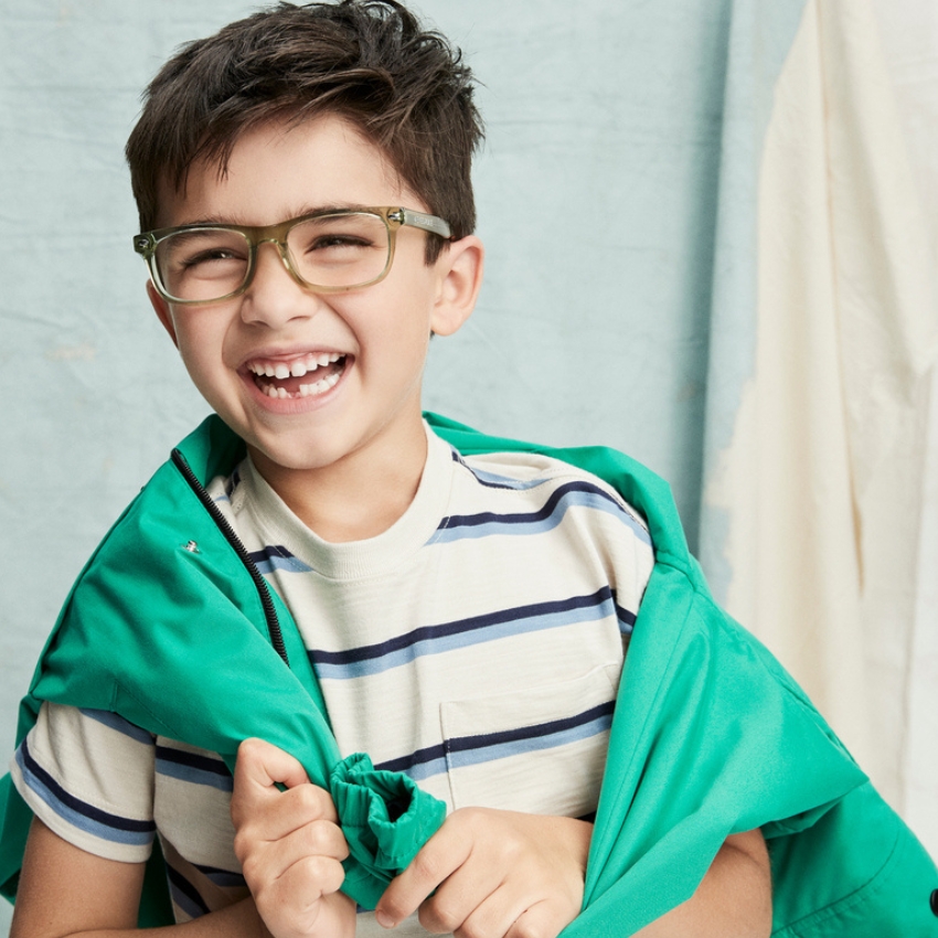 Boy wearing green glasses and a striped shirt, holding a green jacket, smiling widely.