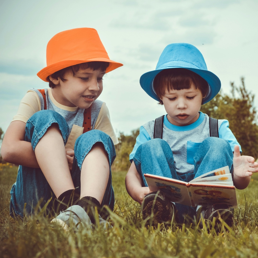 Two children in blue and orange hats sitting on grass, reading a book together.