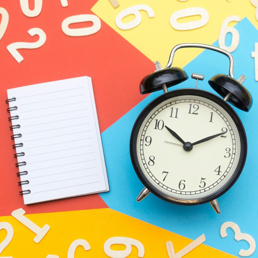 Black analog alarm clock with a white face showing 10:10 next to a small spiral notebook with blank lined pages.