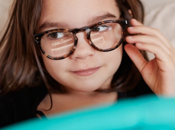Image of a little kid wearing Zenni glasses, with one of her hands touching them, with the reflection of a screen on the lenses.