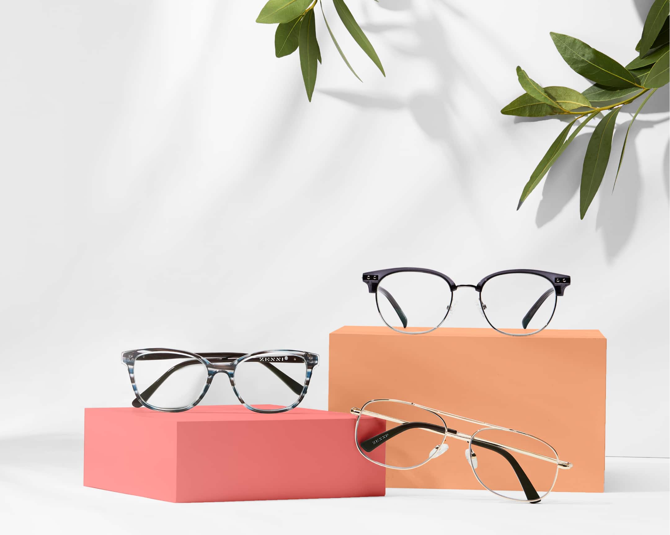 White background and orange risers with 3 prescription glasses under $30: dark grey browline glasses, blue and gray striped rectangle glasses, and gold aviators.